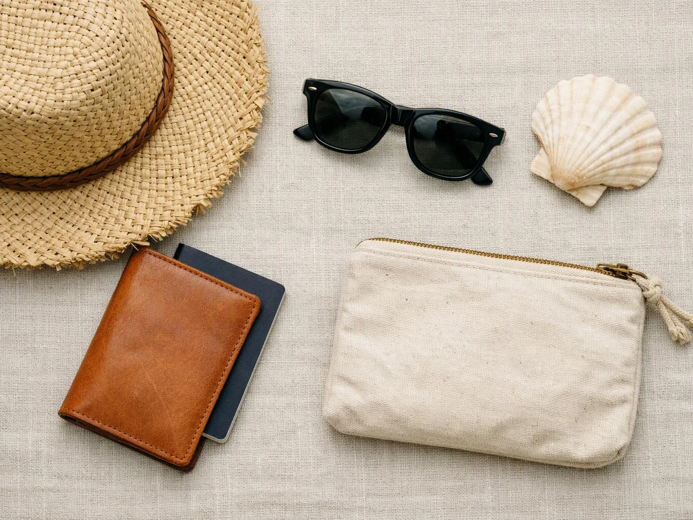 A flat lay shows a straw hat, sunglasses, a shell, a leather wallet, and a canvas pouch on beige fabric