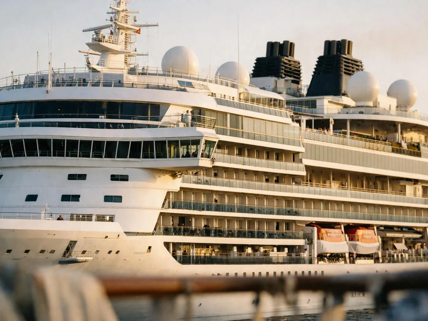 A large white cruise ship sits in calm water, showing stacked decks, windows, radar domes, and black funnels at sunset